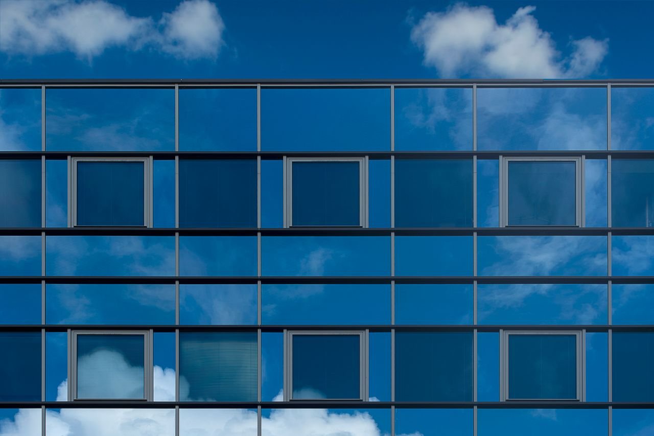 get-in-touch Facade of a modern glass building reflecting the blue sky and clouds.