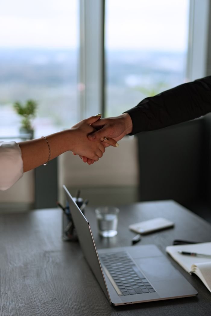 our-story Business professionals shaking hands across a desk in a contemporary office with a laptop.